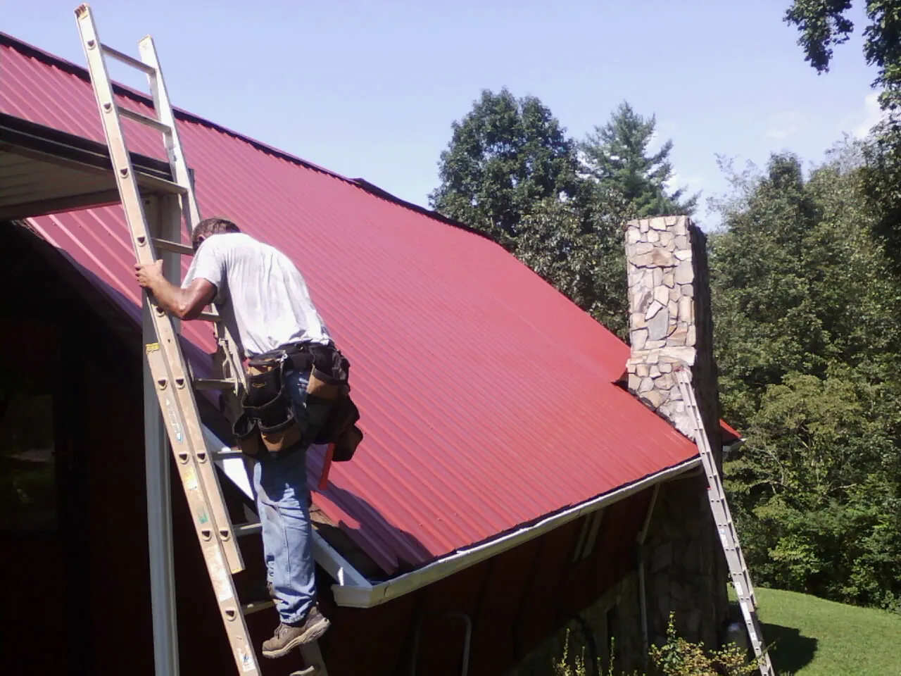 Roofer on a ladder beside a red metal roof and stone chimney — Soffit & Fascia Repair by Roofing Brothers in Meade